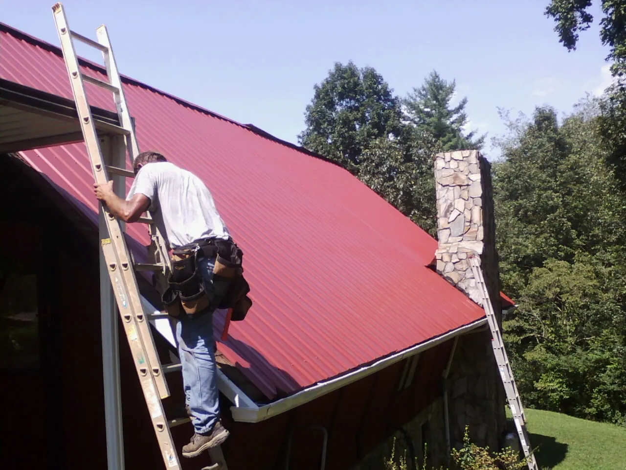 Roofer on a ladder beside a red metal roof and stone chimney — Soffit & Fascia Repair by Roofing Brothers in Nebo